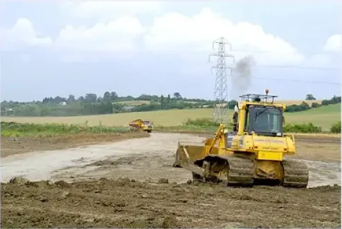 A yellow bulldozer levelling a large earthworks site under pylons, a dump truck in the distance