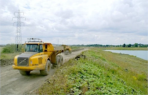 A yellow dump truck on a haul road beside a lake, pylons on the skyline