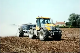 A yellow tractor towing a lime spreader across a ploughed field, lime dust behind it